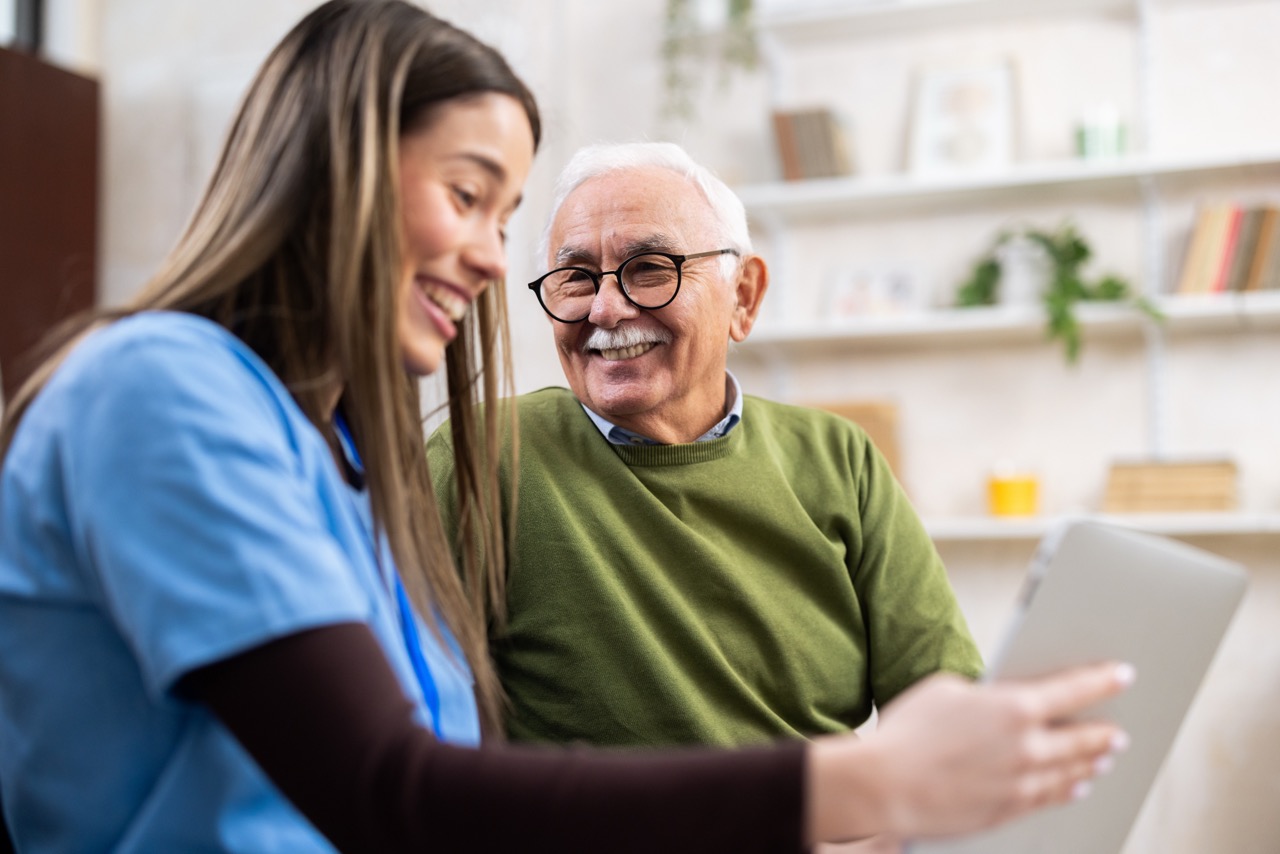 Young caregiver folding laundry while smiling and talking with an elderly woman sitting on a couch at home.